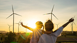 Eine Frau und ein Mädchen, welches getragen wird, blicken gegen die Sonne auf mehrere Windräder. Beide heben je einen Arm seitlich nach oben. Der Blick in den Sonnenuntergang erweckt das Gefühl von Zuversicht. 